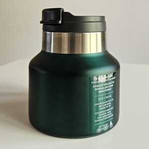 May include: A dark green stainless steel vacuum desktop mug with a black flip lid and a silver rim. A label on the front displays the text: STAINLESS STEEL VACUUM DESKTOP MUG, 20 FL / 591 ML, LEAK RESISTANT FLIP LID, NO SLIDE BASE, TOUGH TO TIP, UP TO 4H.