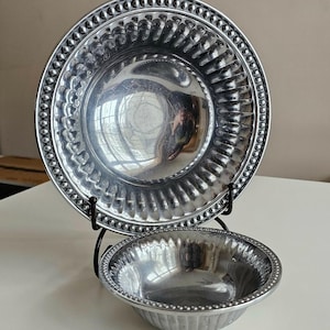 Puede incluir: Dos cuencos de metal plateado, uno grande y otro peque&ntilde;o, expuestos sobre un soporte de metal negro. El cuenco m&aacute;s grande es un plato con un borde de cuentas y un dise&ntilde;o acanalado. El cuenco m&aacute;s peque&ntilde;o tiene un borde de cuentas similar y lados acanalados.