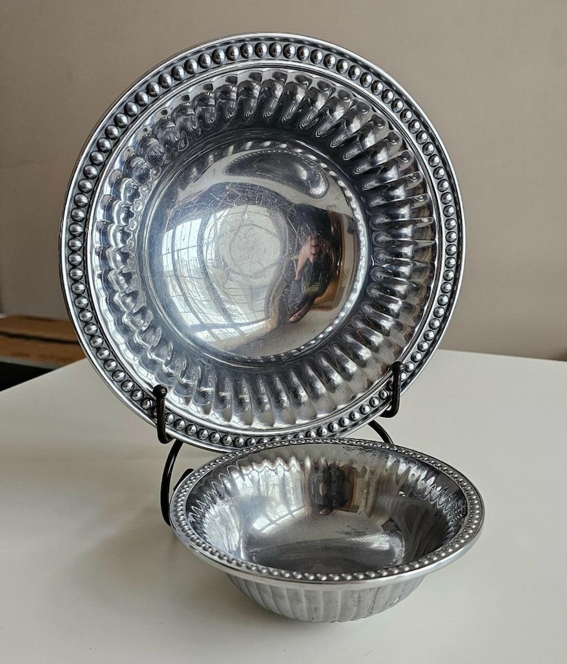 Puede incluir: Dos cuencos de metal plateado, uno grande y otro peque&ntilde;o, expuestos sobre un soporte de metal negro. El cuenco m&aacute;s grande es un plato con un borde de cuentas y un dise&ntilde;o acanalado. El cuenco m&aacute;s peque&ntilde;o tiene un borde de cuentas similar y lados acanalados.