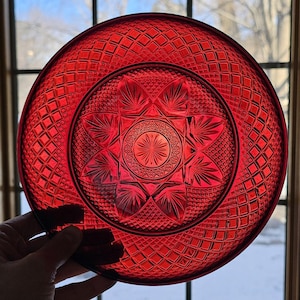 May include: A vibrant red glass plate with a detailed, geometric pattern. The plate features a central starburst design and a diamond-shaped grid pattern around the rim. The plate is held up to the light, showcasing its transparency.