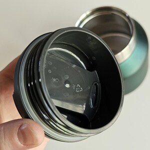 May include: A dark green insulated travel mug with a black plastic lid featuring a sliding closure mechanism. The lid has a circular design with a small vent hole and a recycling symbol with the number 2 and the letters PP molded into the plastic.