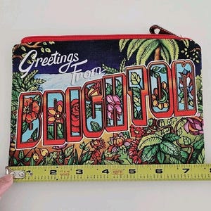 Pu&ograve; includere: Un astuccio rettangolare con cerniera e un design di cartolina colorata. L'astuccio presenta le parole "Greetings From BRIGHTON" in stile vintage, con illustrazioni floreali e di palme. L'astuccio misura circa 20 cm di larghezza.