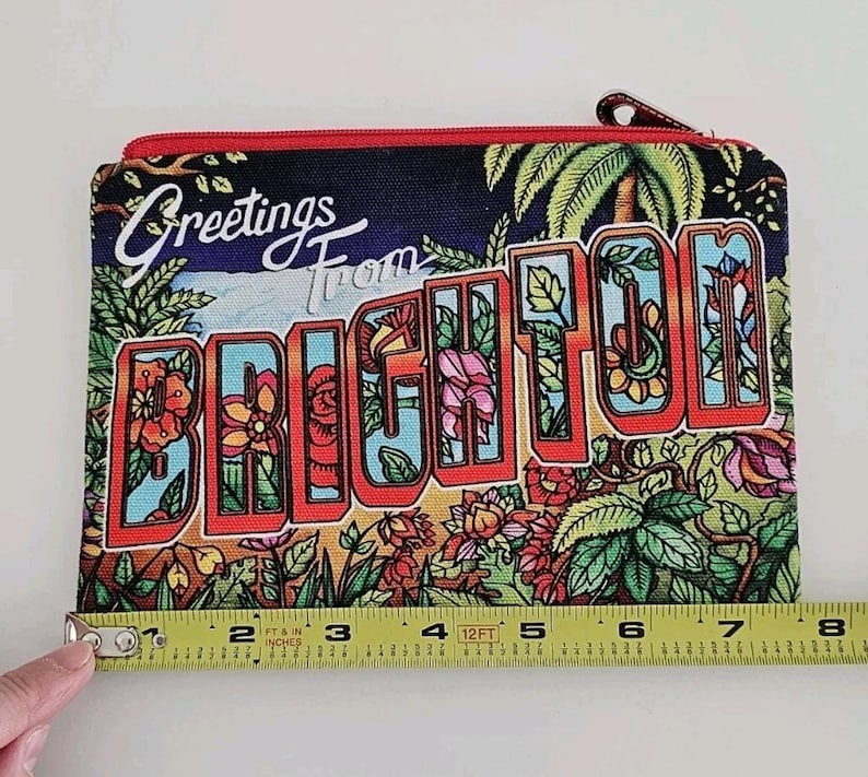 Pu&ograve; includere: Un astuccio rettangolare con cerniera e un design di cartolina colorata. L'astuccio presenta le parole "Greetings From BRIGHTON" in stile vintage, con illustrazioni floreali e di palme. L'astuccio misura circa 20 cm di larghezza.