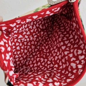 Pu&ograve; includere: Vista aperta di una piccola pochette rossa con cerniera e fodera a cuori bianchi. L'esterno presenta un motivo floreale e un tirazip argentato. L'interno &egrave; rosso vivo con un motivo ripetuto di cuori bianchi.