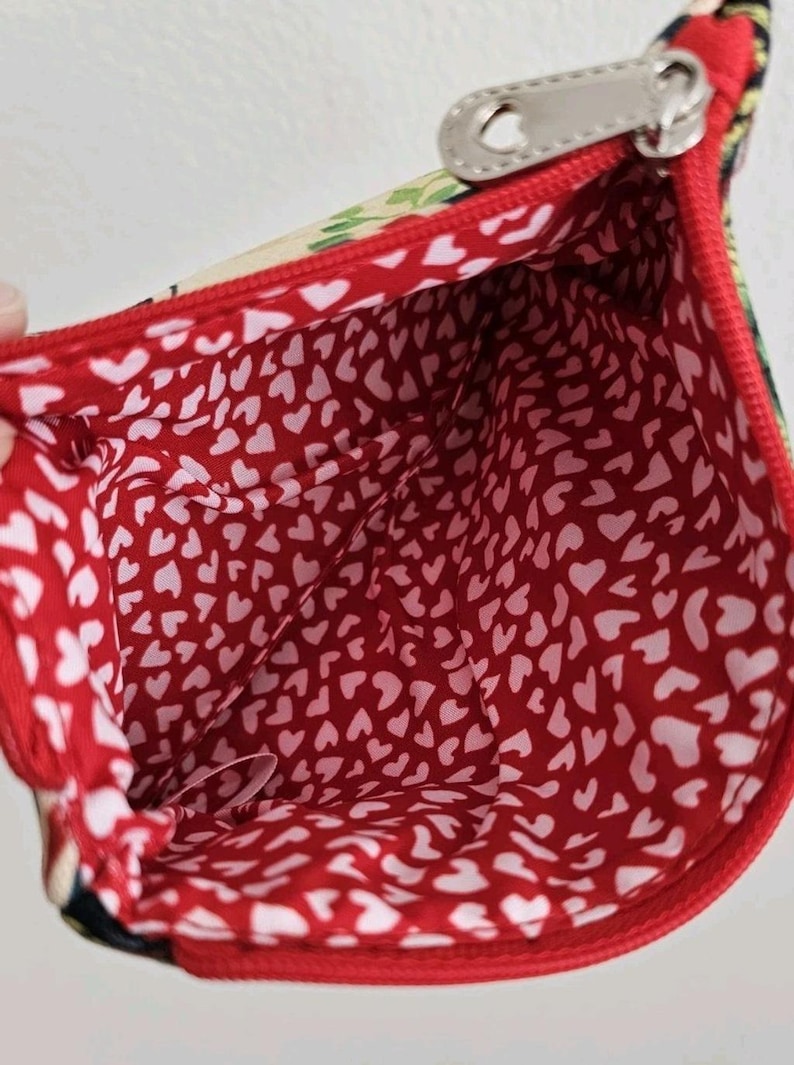 Pu&ograve; includere: Vista aperta di una piccola pochette rossa con cerniera e fodera a cuori bianchi. L'esterno presenta un motivo floreale e un tirazip argentato. L'interno &egrave; rosso vivo con un motivo ripetuto di cuori bianchi.