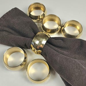 May include: A set of six gold-toned metal napkin rings with a smooth, polished finish, except for one ring that features a distinct hammered texture. One of the hammered napkin rings is shown holding a folded dark brown fabric napkin.