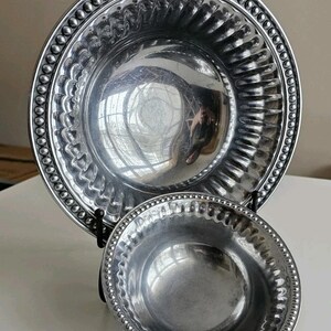 Puede incluir: Dos cuencos de metal plateado. El cuenco m&aacute;s grande tiene un borde ancho con borde de cuentas y un interior acanalado. El cuenco m&aacute;s peque&ntilde;o tambi&eacute;n tiene un borde de cuentas. Ambos cuencos se exhiben en un soporte negro.