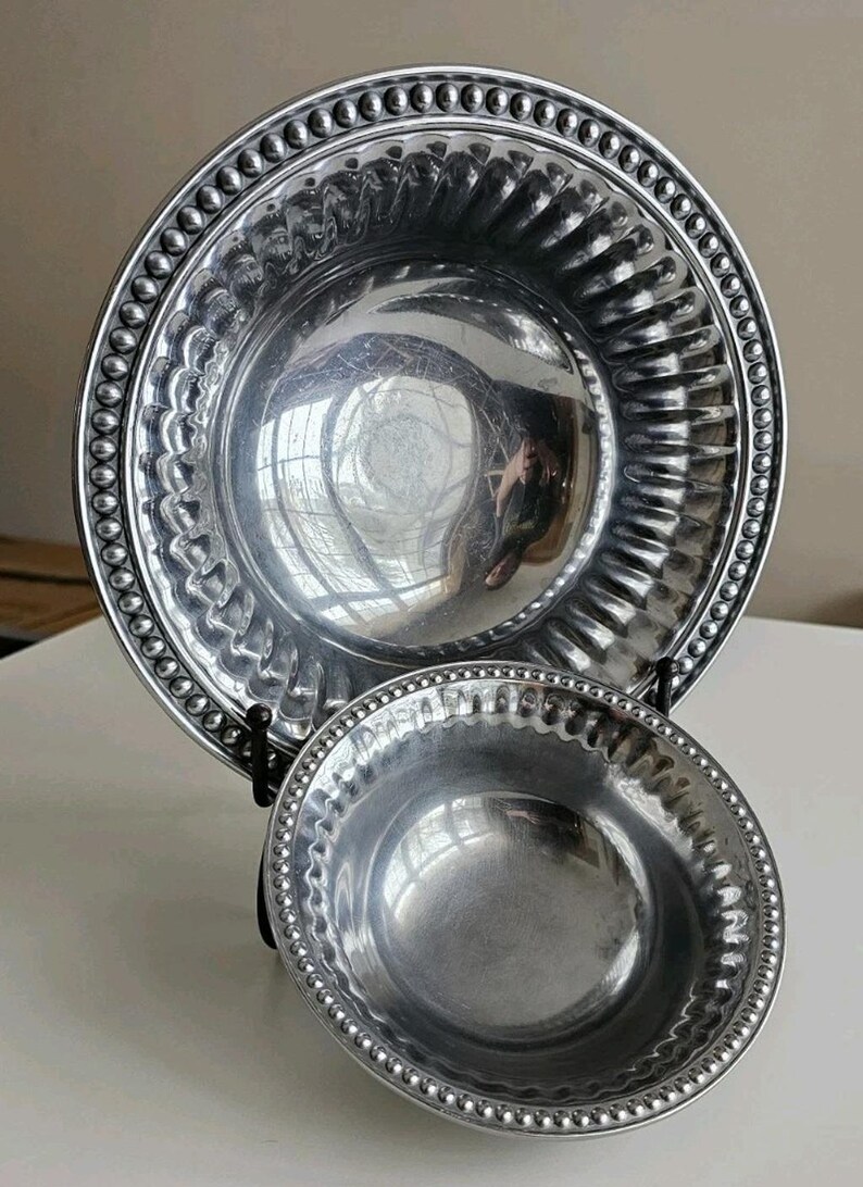 Puede incluir: Dos cuencos de metal plateado. El cuenco m&aacute;s grande tiene un borde ancho con borde de cuentas y un interior acanalado. El cuenco m&aacute;s peque&ntilde;o tambi&eacute;n tiene un borde de cuentas. Ambos cuencos se exhiben en un soporte negro.