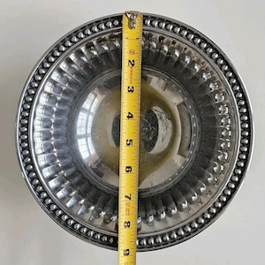 Puede incluir: Un cuenco de metal plateado con un borde con cuentas y un interior acanalado. El cuenco mide aproximadamente 23 cm de di&aacute;metro, medido con una cinta m&eacute;trica amarilla. El cuenco es redondo y reflectante.