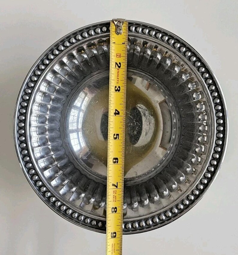Puede incluir: Un cuenco de metal plateado con un borde con cuentas y un interior acanalado. El cuenco mide aproximadamente 23 cm de di&aacute;metro, medido con una cinta m&eacute;trica amarilla. El cuenco es redondo y reflectante.