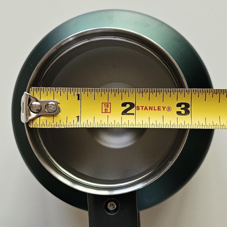 May include: A dark green circular container with a metallic interior, shown from a top-down perspective. A yellow Stanley measuring tape is positioned across the opening, indicating an internal diameter of approximately 3.25 inches.