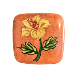 May include: Square orange decorative box with a hand-painted yellow flower design. The flower has red and yellow details, with green leaves and stem. The box has a textured surface.