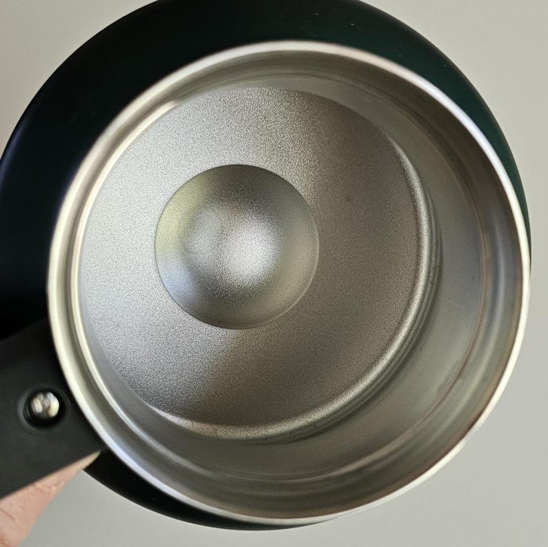 May include: A top-down view of a dark-colored mug with a metallic silver interior. The mug features a smooth, matte black exterior finish and a sturdy black handle attached with a visible silver screw. The interior surface has a textured finish.