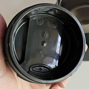 May include: A black plastic circular lid for a travel mug featuring a central raised rectangular section with a small sip opening. The underside displays molded markings including a recycling symbol with PP, the number 2, and a circular date dial.
