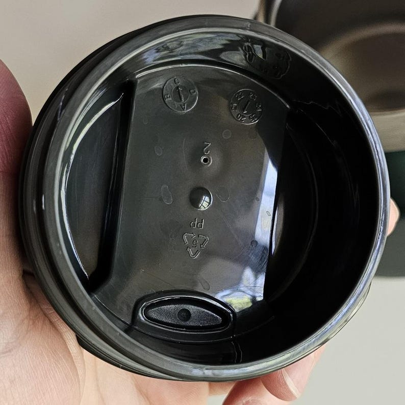 May include: A black plastic circular lid for a travel mug featuring a central raised rectangular section with a small sip opening. The underside displays molded markings including a recycling symbol with PP, the number 2, and a circular date dial.