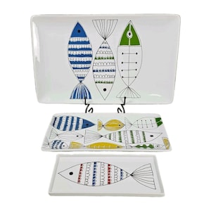 May include: Three rectangular white ceramic serving platters of varying sizes, each decorated with stylized fish illustrations in blue, green, yellow, and red. The designs feature linear patterns, dots, and stripes on the fish bodies.
