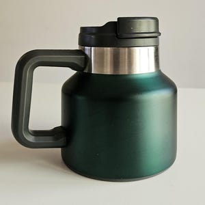 May include: A dark forest green insulated mug with a wide, cylindrical body and a matte finish. It features a sturdy black plastic handle on the side and a matching black plastic lid with a sliding closure mechanism. The upper rim is stainless steel.