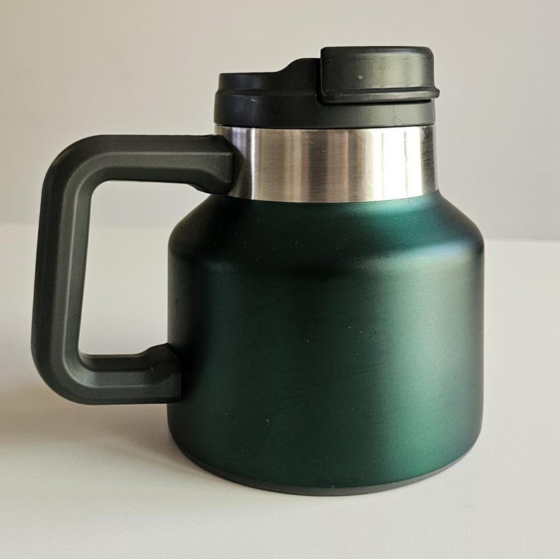 May include: A dark forest green insulated mug with a wide, cylindrical body and a matte finish. It features a sturdy black plastic handle on the side and a matching black plastic lid with a sliding closure mechanism. The upper rim is stainless steel.