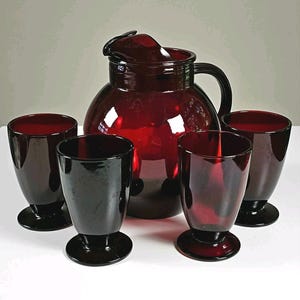 May include: A deep red glass pitcher with a matching lid and handle, accompanied by four matching footed drinking glasses. The set features a smooth, translucent finish and a rounded silhouette, creating a cohesive collection of dark ruby-colored glassware.