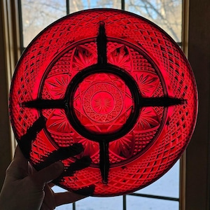 May include: A vibrant red glass serving dish with a diamond and starburst pattern. The dish has a circular shape with a black cross-shaped divider. The center features a star design. The dish is held up in front of a window.