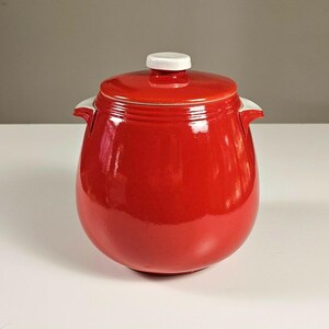 May include: A glossy, bright red ceramic jar with a rounded, bulbous body and two small side handles. The jar features a matching red lid topped with a small, circular white knob. The surface has a smooth, reflective finish throughout.