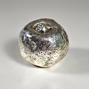 May include: A small, round, silver-colored decorative object shaped like a piece of fruit. The surface has a highly reflective, metallic finish with a textured, pitted appearance and a small, raised protrusion at the top center.