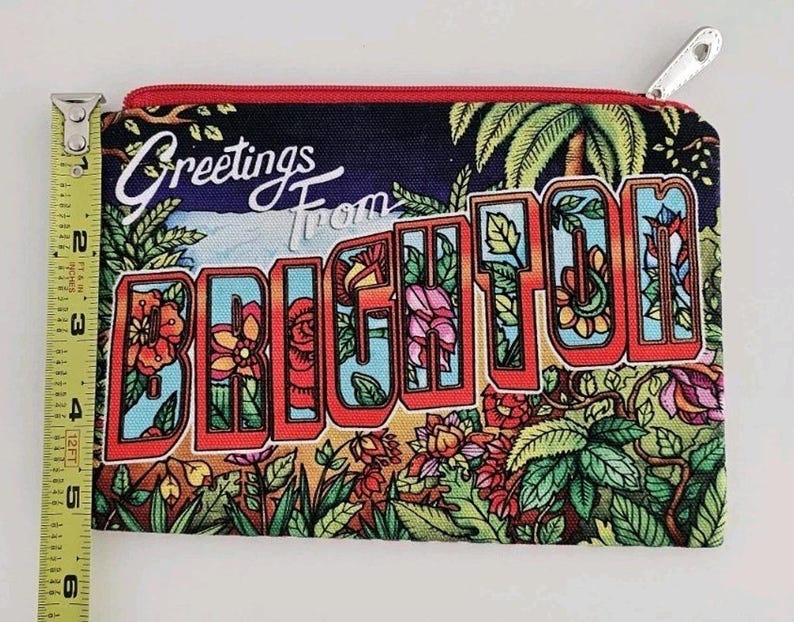 Pu&ograve; includere: Un astuccio rettangolare con cerniera souvenir di Brighton, largo circa 15 cm, con cerniera rossa e tiretto argentato. L'astuccio presenta un colorato design da cartolina con le parole "Greetings From Brighton" e illustrazioni floreali e di palme.