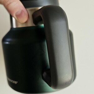 May include: A dark forest green insulated tumbler featuring a matte finish, a sturdy black side handle, and a metallic silver rim near the top. The base of the tumbler displays the partial white text logo for Starbucks on the lower side surface.
