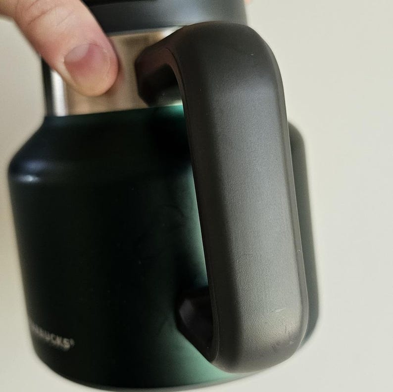 May include: A dark forest green insulated tumbler featuring a matte finish, a sturdy black side handle, and a metallic silver rim near the top. The base of the tumbler displays the partial white text logo for Starbucks on the lower side surface.