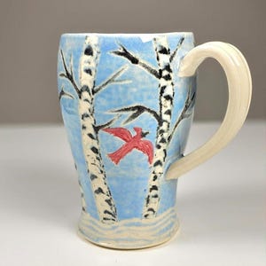 May include: A ceramic mug with a light blue background featuring a painted scene of two white birch trees with black markings and a small red bird in flight. The mug has a smooth, cream-colored handle and a base with subtle white wavy lines.
