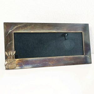 May include: A rectangular metal picture frame with a dark, burnished finish and a black velvet interior. The bottom left corner features a raised brass-toned relief of a fish jumping out of a basket surrounded by three stalks of wheat.