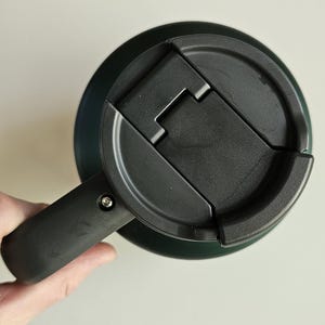 May include: A dark green travel mug featuring a black plastic lid with a hinged flip-top closure. The mug includes a sturdy black handle attached to the side with a visible silver screw, providing a secure grip for the cylindrical container.