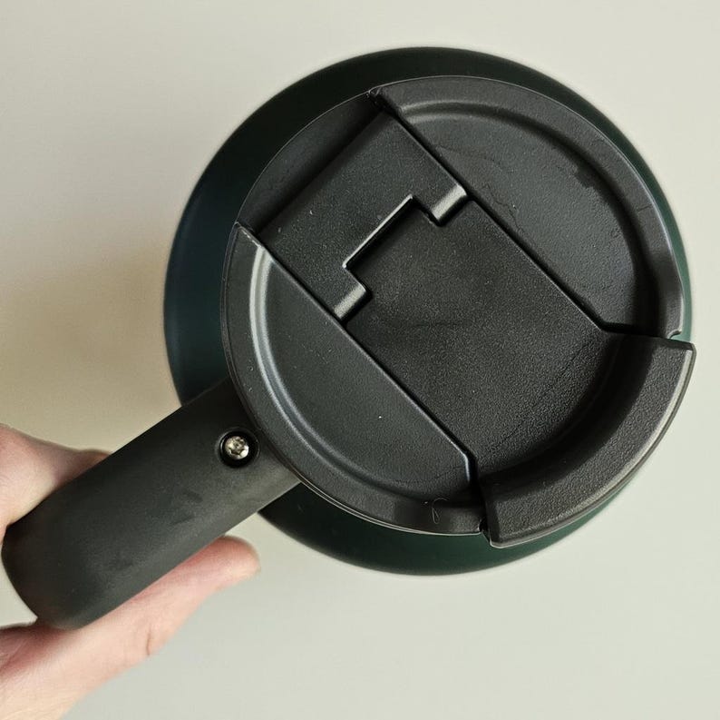 May include: A dark green travel mug featuring a black plastic lid with a hinged flip-top closure. The mug includes a sturdy black handle attached to the side with a visible silver screw, providing a secure grip for the cylindrical container.