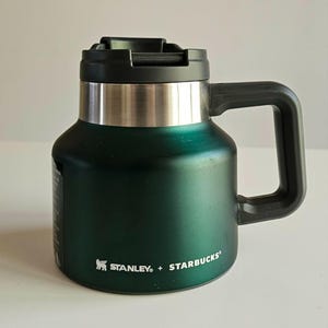 May include: A dark green Stanley + Starbucks insulated mug with a wide base, a metallic silver band near the top, and a black lid with a handle. The text Stanley + Starbucks is printed in white on the lower front of the metallic green body.