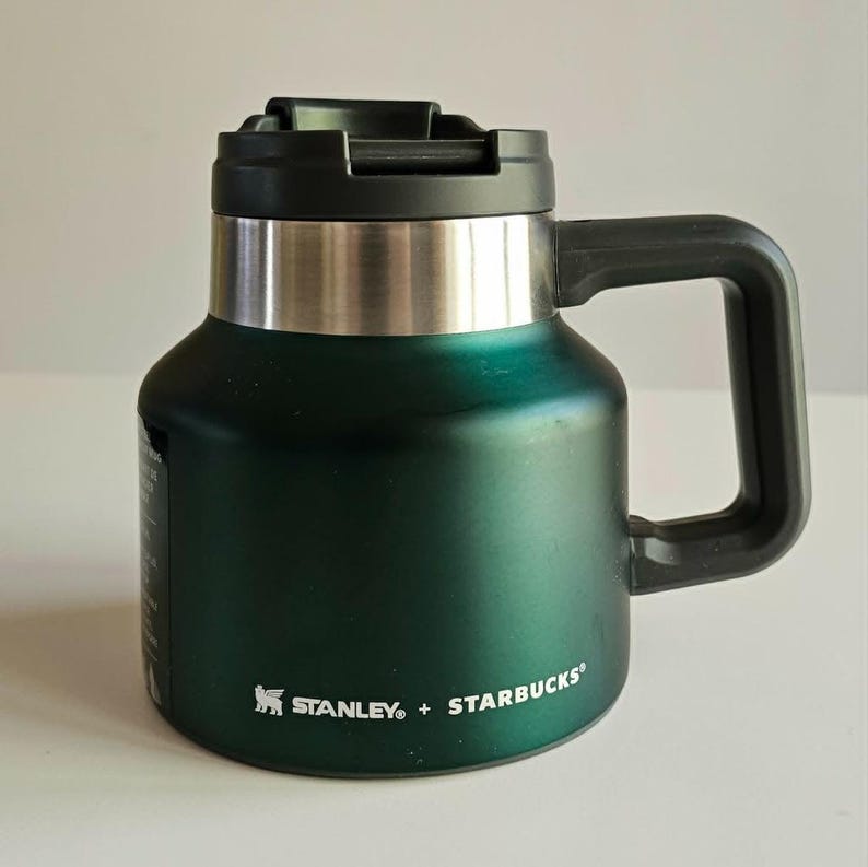 May include: A dark green Stanley + Starbucks insulated mug with a wide base, a metallic silver band near the top, and a black lid with a handle. The text Stanley + Starbucks is printed in white on the lower front of the metallic green body.