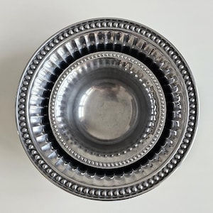 Puede incluir: Tres cuencos met&aacute;licos plateados anidados. El cuenco m&aacute;s grande tiene un borde con cuentas, el cuenco del medio tiene un dise&ntilde;o acanalado y el cuenco m&aacute;s peque&ntilde;o es liso. Los cuencos est&aacute;n dispuestos sobre una superficie blanca.