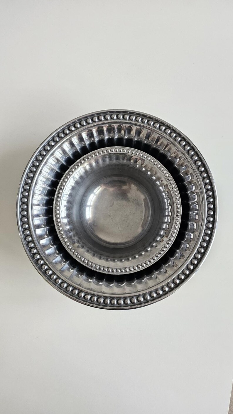 Puede incluir: Tres cuencos met&aacute;licos plateados anidados. El cuenco m&aacute;s grande tiene un borde con cuentas, el cuenco del medio tiene un dise&ntilde;o acanalado y el cuenco m&aacute;s peque&ntilde;o es liso. Los cuencos est&aacute;n dispuestos sobre una superficie blanca.