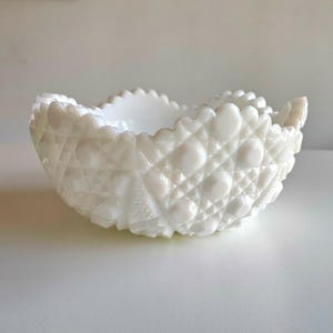 May include: A white, vintage-style bowl with a textured surface. The bowl features a diamond and dot pattern, with a scalloped rim. The bowl is made of opaque white glass and is likely used for serving or display.