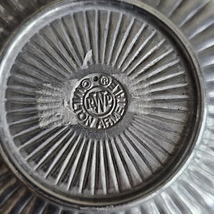 Puede incluir: Primer plano de un plato de peltre con borde acanalado y un emblema central circular. El emblema presenta el texto "WILTON ARMETALE" alrededor de un dise&ntilde;o central. La superficie del plato tiene un aspecto met&aacute;lico texturizado.