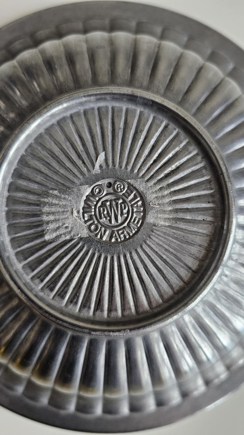 Puede incluir: Primer plano de un plato de peltre con borde acanalado y un emblema central circular. El emblema presenta el texto "WILTON ARMETALE" alrededor de un dise&ntilde;o central. La superficie del plato tiene un aspecto met&aacute;lico texturizado.