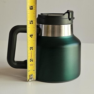 May include: A dark green insulated mug with a black handle and a black lid. A yellow Stanley measuring tape is positioned vertically against the mug, showing a height measurement of approximately 6 inches from the base to the top of the lid.