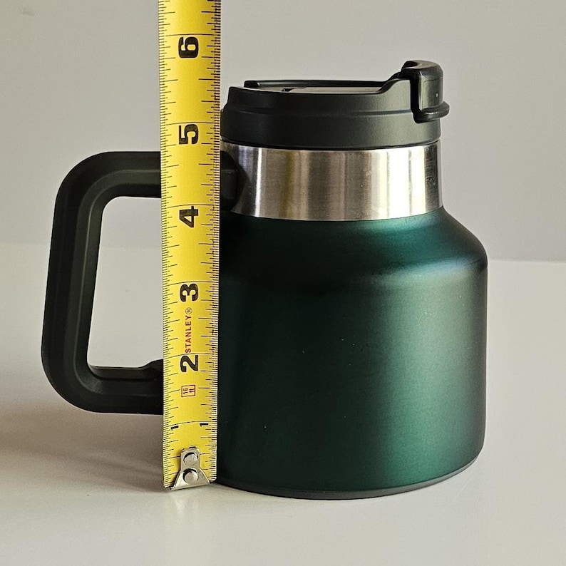 May include: A dark green insulated mug with a black handle and a black lid. A yellow Stanley measuring tape is positioned vertically against the mug, showing a height measurement of approximately 6 inches from the base to the top of the lid.