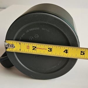 May include: A dark gray mug with a black base featuring a yellow Stanley measuring tape across the bottom. The tape shows a diameter of approximately 3.5 inches. The base includes molded text reading BPA FREE, DISHWASHER SAFE, and IN CHINA.