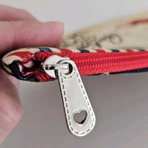 Pu&ograve; includere: Primo piano di una piccola pochette con cerniera rossa. Il tirazip &egrave; color argento con un ritaglio a forma di cuore. Il tessuto della pochette presenta un mix di colori e motivi.