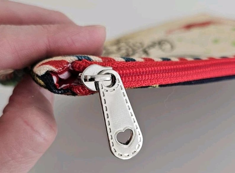 Pu&ograve; includere: Primo piano di una piccola pochette con cerniera rossa. Il tirazip &egrave; color argento con un ritaglio a forma di cuore. Il tessuto della pochette presenta un mix di colori e motivi.