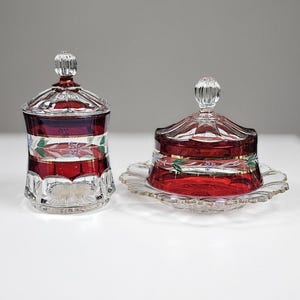 May include: Two pieces of ruby red and clear cut glass tableware with lids. Each piece features a horizontal band of red glass, gold trim, and hand-painted floral motifs with green leaves and small blue flowers on a clear glass base.