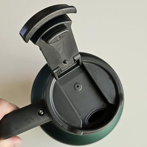 May include: A dark green travel mug with a black plastic lid featuring an open flip-top mechanism. The lid has a circular vent hole and a contoured drinking spout. The mug includes a black handle attached to the side with a visible metal screw.