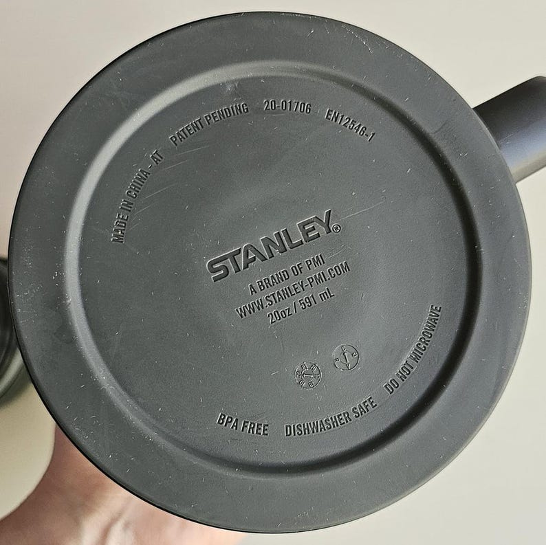 May include: The circular black base of a Stanley mug features embossed text including STANLEY, A BRAND OF PMI, WWW.STANLEY-PMI.COM, 20oz / 591 mL, MADE IN CHINA - AT, PATENT PENDING, 20-01706, EN12546-1, BPA FREE, DISHWASHER SAFE, and DO NOT MICROWAVE.