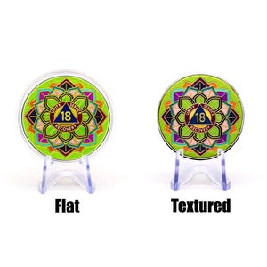 May include: Two round, green and gold medallions with a colorful geometric design and the words "Unity, Service, Recovery" and the number "18". One medallion is labeled "Flat" and the other "Textured". Both are displayed on clear stands.