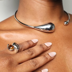 Op de afbeelding: Een zilverkleurige ketting met een druppelvormige hanger en een bijpassende ring met een bruine steen. De sieraden worden getoond op de borst en hand van een persoon, waarbij het elegante ontwerp en de metallic glans worden benadrukt.